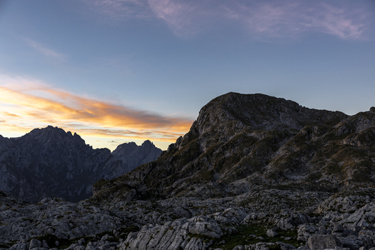 View of rugged mountain peaks rise against a soft dawn sky with hues of orange and pink painting the horizon contrasting with the dark rocky terrain Picos de Europa Asturias Spain.