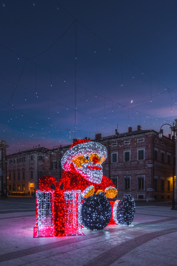View of a large illuminated Santa Claus figure with a gift box in a town square decorated with festive string lights at dusk in Senigallia Marche Italy.
