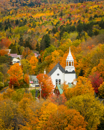 Aerial view of fall foliage in a small Vermont town featuring a white church with a steeple in vibrant autumn trees. Island Pond Brighton Vermont USA. Aerial view of fall foliage in a small Vermont town featuring a white church with a steeple in vibrant autumn trees. Island Pond Brighton Vermont USA.