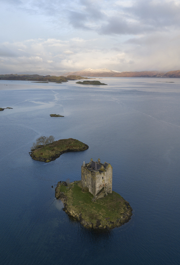 Aerial view of Castle Stalker on a small tidal islet surrounded by the calm blue waters of Loch Laich with distant mountains under a cloudy sky in Appin Scotland United Kingdom. Aerial view of Castle Stalker on a small tidal islet surrounded by the calm blue waters of Loch Laich with distant mountains under a cloudy sky in Appin Scotland United Kingdom.
