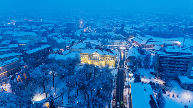Aerial view of the snow covered town center featuring illuminated historic buildings a roundabout and snow dusted trees under a blue twilight sky in Sremska Mitrovica Vojvodina Serbia.