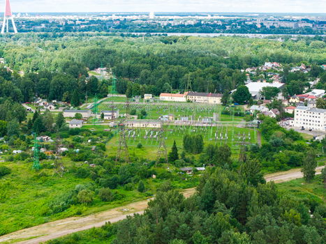 Aerial view of an electrical substation surrounded by lush green forest and residential buildings with the Riga TV Tower visible in the distance in Riga Latvia.