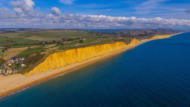 Aerial view of the iconic golden sandstone cliffs of West Bay stretching along the coastline with a sandy beach and rolling green fields in West Bay England United Kingdom. Aerial view of the iconic golden sandstone cliffs of West Bay stretching along the coastline with a sandy beach and rolling green fields in West Bay England United Kingdom.