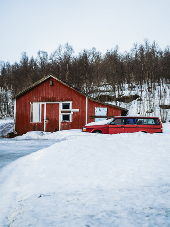 View of a vibrant red vintage car parked in deep snow near a rustic red wooden building creating a striking contrast against the white landscape Bardufoss Norway. View of a vibrant red vintage car parked in deep snow near a rustic red wooden building creating a striking contrast against the white landscape Bardufoss Norway.