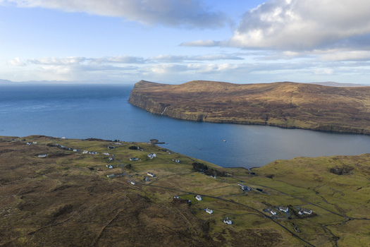 Aerial view of the coastal village of Milovaig with scattered white houses on green hills overlooking the blue waters of Loch Pooltiel and rugged cliffs Milovaig Scotland United Kingdom.