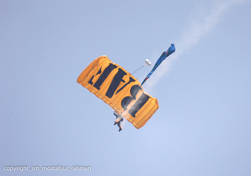 Dhaka Bangladesh 25 March 2026 View of a parachutist descending with a canopy of vibrant orange emblazoned with bold dark lettering against the clear sky.