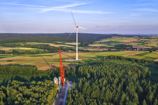 Aerial view of a wind turbine under construction with a large orange crane positioned near a dense forest and agricultural fields in Hofgeismar Hessen Germany.