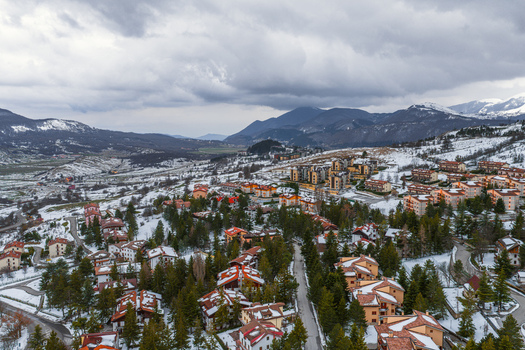 Aerial view of Pescocostanzo featuring snow covered rooftops dense pine trees and mountain slopes under a dramatic cloudy sky in Pescocostanzo Abruzzo Italy.