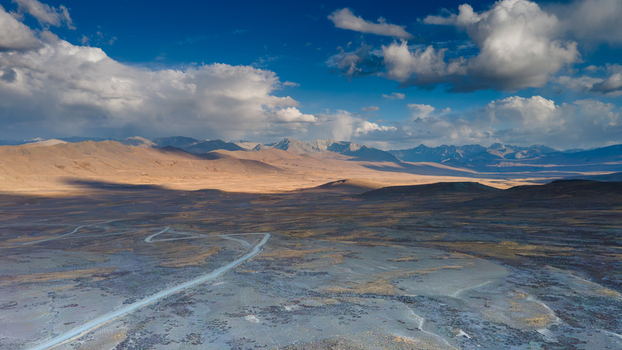 Aerial view of Deosai National Park featuring vast high altitude plains winding dirt roads and distant snow capped mountains under a dramatic cloudy sky in Skardu Gilgit Pakistan.