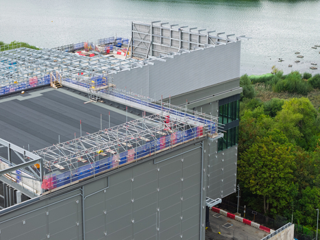 Aerial view of a data center under construction with scaffolding and metal framework on the roof situated near a body of water in the United Kingdom.