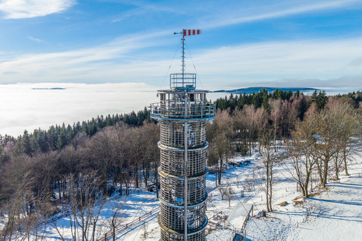 Aerial view of the Majak Jary Cimrmana observation tower surrounded by snow covered forest and a sea of clouds under a clear blue sky in Korenov Liberecky kraj Czechia. Aerial view of the Majak Jary Cimrmana observation tower surrounded by snow covered forest and a sea of clouds under a clear blue sky in Korenov Liberecky kraj Czechia.
