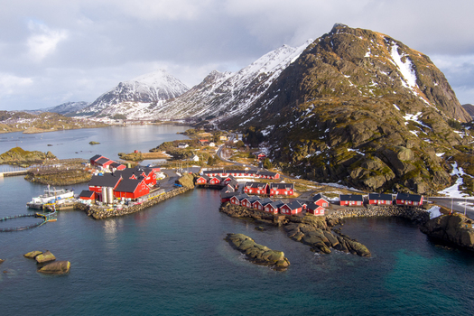 Aerial view of the fishing village of Lofoten with its iconic red wooden cabins fish farming pens and snow capped mountains in the background Nordland Norway.