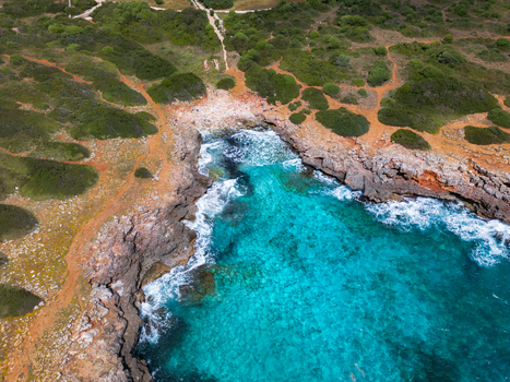 Aerial view of the rocky coastline and turquoise waters of a secluded cove surrounded by green Mediterranean vegetation and dirt paths in Sa Coma Balearic Islands Spain. Aerial view of the rocky coastline and turquoise waters of a secluded cove surrounded by green Mediterranean vegetation and dirt paths in Sa Coma Balearic Islands Spain.