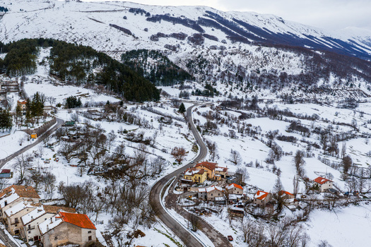 Aerial view of Pescocostanzo village nestled in snow covered mountains featuring winding roads stone buildings with orange rooftops and pine forests in Pescocostanzo Abruzzo Italy.