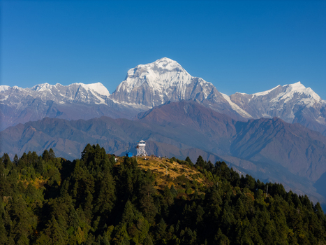Aerial view of the majestic Dhaulagiri mountain range with snow capped peaks rising above a forested ridge and a small observation structure in Nepal.