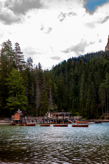 View of the wooden boathouse and rowing boats on the calm waters of Lago di Braies in the Dolomites surrounded by dense pine forests under a cloudy sky in Rocca Pietore Veneto Italy. View of the wooden boathouse and rowing boats on the calm waters of Lago di Braies in the Dolomites surrounded by dense pine forests under a cloudy sky in Rocca Pietore Veneto Italy.