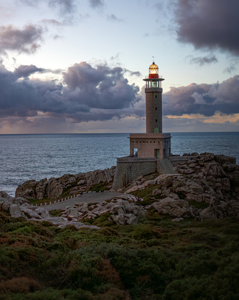 View of Faro de Punta Nariga lighthouse standing on a rugged rocky cliff overlooking the Atlantic Ocean under a dramatic cloudy sky at twilight in Malpica A Coruna Spain. View of Faro de Punta Nariga lighthouse standing on a rugged rocky cliff overlooking the Atlantic Ocean under a dramatic cloudy sky at twilight in Malpica A Coruna Spain.