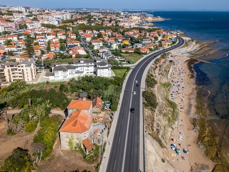 Aerial view of Avenida Marginal winding along the coastline with a sandy beach residential houses and cars driving on the road in Estoril Lisboa Portugal.