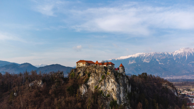 Aerial view of Bled Castle perched on a steep rocky cliff with the snow capped Julian Alps in the background under a blue sky in Bled Radovljica Slovenia.