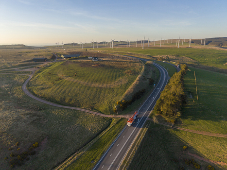 Aerial view of a red truck traveling along a winding road through lush green fields and a vast wind farm under a golden sunset sky. Aerial view of a red truck traveling along a winding road through lush green fields and a vast wind farm under a golden sunset sky.