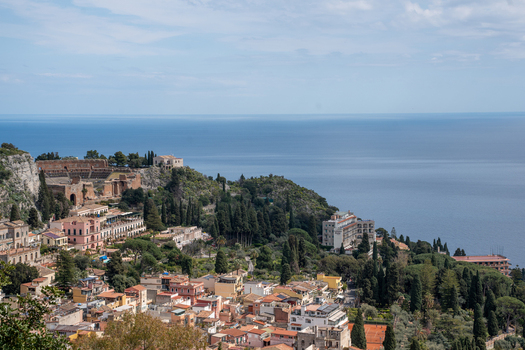 View of ancient ruins cascading down to the turquoise sea with a charming town nestled amid lush greenery under a vast sky Taormina Sicily Italy.