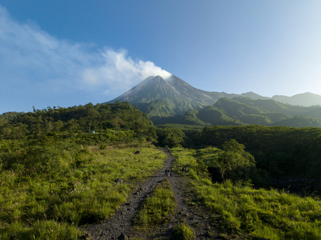 Aerial view of Bunker Kaliadem Merapi and the smoking crater of Mount Merapi volcano with lush green hills and a dirt path under a blue sky Kecamatan Pakem Daerah Istimewa Yogya Indonesia. Aerial view of Bunker Kaliadem Merapi and the smoking crater of Mount Merapi volcano with lush green hills and a dirt path under a blue sky Kecamatan Pakem Daerah Istimewa Yogya Indonesia.