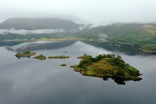 Aerial view of small islands in a calm loch surrounded by misty mountains and lush greenery in Glencoe Scotland United Kingdom.