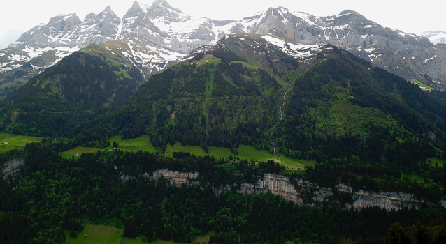 Aerial view of lush green alpine slopes dense pine forests and rocky cliffs under snow capped mountain peaks in Champery Valais Switzerland. Aerial view of lush green alpine slopes dense pine forests and rocky cliffs under snow capped mountain peaks in Champery Valais Switzerland.