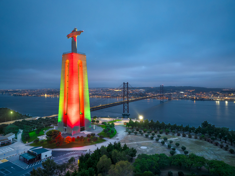 Aerial view of the Sanctuary of Christ the King illuminated in red and green lights with the 25 de Abril Bridge and Tagus River at dusk Almada Setubal Portugal. Aerial view of the Sanctuary of Christ the King illuminated in red and green lights with the 25 de Abril Bridge and Tagus River at dusk Almada Setubal Portugal.