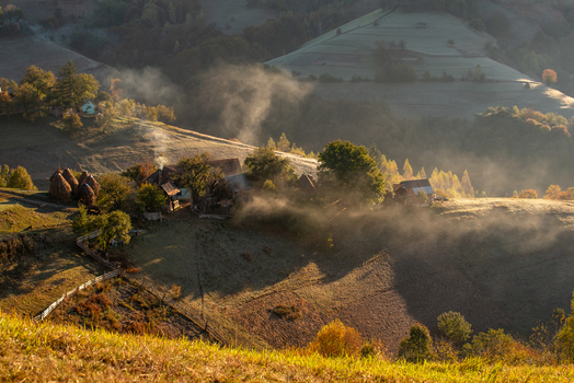 View of a traditional rural landscape with morning mist rising over wooden houses haystacks and rolling hills in the autumn light Pestera Brasov Romania. View of a traditional rural landscape with morning mist rising over wooden houses haystacks and rolling hills in the autumn light Pestera Brasov Romania.