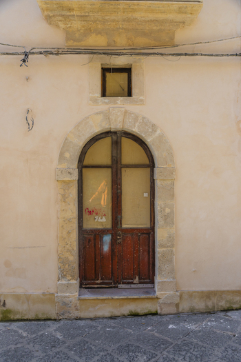 View of an old wooden door with a stone arch and a small window above it set in a peach colored wall with visible wires Ortigia Sicily Italy.
