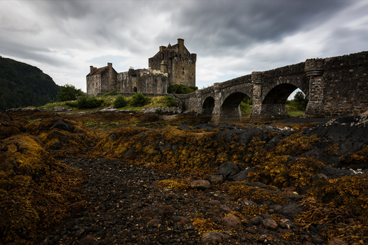 View of Eilean Donan Castle with its iconic stone bridge and rocky shore under a dramatic cloudy sky Scotland.
