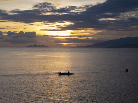 Aerial view of a lone fisherman in a small wooden boat on the calm sea at sunset with distant mountains in Flores Indonesia.