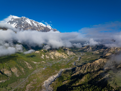 Aerial view of the snow capped Himalayan peaks rising above a lush green valley with a riverbed and a small airstrip in Nepal.