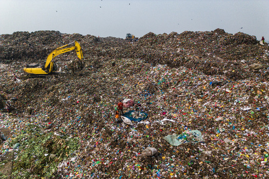 Aminbazar Bangladesh 26 January 2025 Aerial view of the Aminbazar landfill with a yellow excavator moving waste and people scavenging through piles of garbage under a hazy sky. Aminbazar Bangladesh 26 January 2025 Aerial view of the Aminbazar landfill with a yellow excavator moving waste and people scavenging through piles of garbage under a hazy sky.