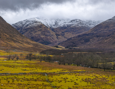 Aerial view of snow capped mountains overlooking a vast valley with golden fields and bare trees under a cloudy sky in Scotland United Kingdom. Aerial view of snow capped mountains overlooking a vast valley with golden fields and bare trees under a cloudy sky in Scotland United Kingdom.