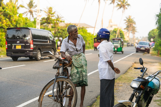 Sri Lanka 21 January 2026 View of two local men talking on the side of a busy road with a bicycle traffic and palm trees in the background.