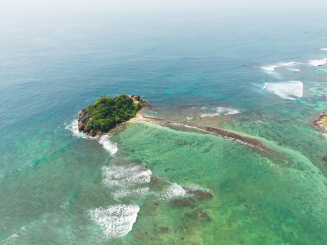 Aerial view of Devil s Rock a small island covered in green vegetation surrounded by turquoise ocean waves and a misty horizon in Ahangama Southern Province Sri Lanka. Aerial view of Devil s Rock a small island covered in green vegetation surrounded by turquoise ocean waves and a misty horizon in Ahangama Southern Province Sri Lanka.