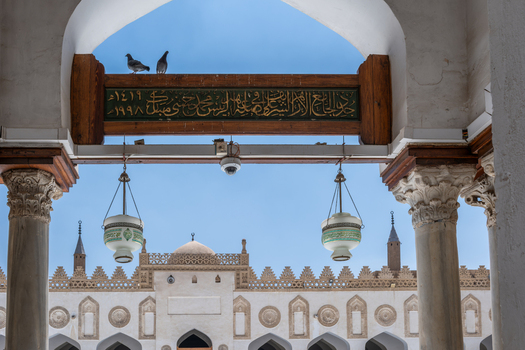 View of the historic Al Azhar Mosque with its ornate white arches hanging lanterns and pigeons perched on a wooden beam under a clear blue sky Cairo Egypt. View of the historic Al Azhar Mosque with its ornate white arches hanging lanterns and pigeons perched on a wooden beam under a clear blue sky Cairo Egypt.