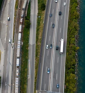 Aerial view of a train on tracks running parallel to a highway with cars and a truck situated along the waterfront in Melide Ticino Switzerland. Aerial view of a train on tracks running parallel to a highway with cars and a truck situated along the waterfront in Melide Ticino Switzerland.