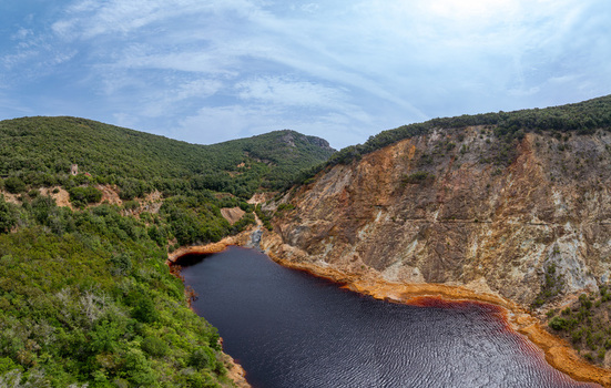 Aerial view of the Laghetto delle conche a striking red hued mining lake surrounded by lush green hills and steep rocky cliffs in Rio Marina Toscana Italy.