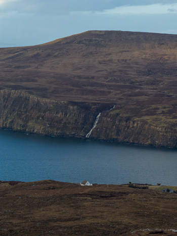 Aerial view of the rugged cliffs and a cascading waterfall flowing into the blue sea near a white cottage in Milovaig Scotland United Kingdom.