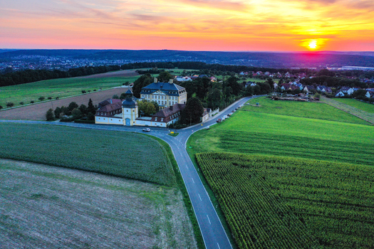 Aerial view of Schloss Jagersburg surrounded by lush green fields and a winding road under a vibrant sunset sky Eggolsheim Bayern Germany.