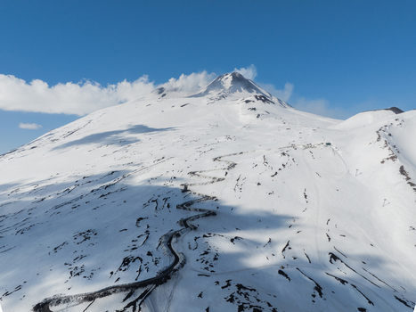 Aerial view of the snow covered south slope of Mount Etna with a winding mountain road and volcanic peak under a clear blue sky in Nicolosi Sicilia Italy. Aerial view of the snow covered south slope of Mount Etna with a winding mountain road and volcanic peak under a clear blue sky in Nicolosi Sicilia Italy.