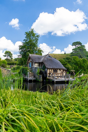 View of a quaint thatched roof cottage on a tranquil pond amidst vibrant green foliage under a bright sky Hobbiton Movie Set Waikato Region New Zealand. View of a quaint thatched roof cottage on a tranquil pond amidst vibrant green foliage under a bright sky Hobbiton Movie Set Waikato Region New Zealand.