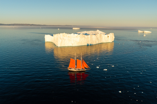 Aerial view of a sailboat with vibrant red sails navigating through deep blue waters near a massive white iceberg under the soft light of the golden hour in Greenland. Aerial view of a sailboat with vibrant red sails navigating through deep blue waters near a massive white iceberg under the soft light of the golden hour in Greenland.
