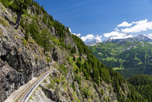 View of the VerticAlp Emosson railway track winding along the steep rocky mountainside with pine forests and distant peaks in Le Chatelard Fribourg Switzerland. View of the VerticAlp Emosson railway track winding along the steep rocky mountainside with pine forests and distant peaks in Le Chatelard Fribourg Switzerland.