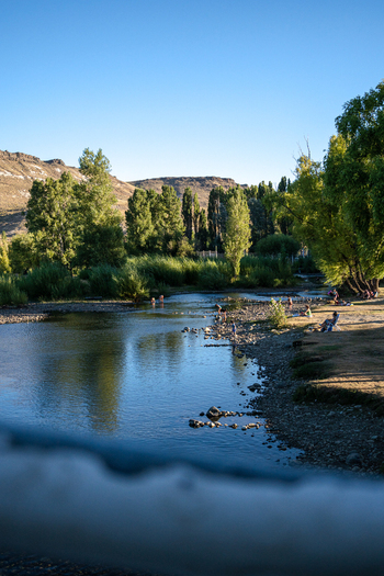 View of people relaxing on the banks of the Rio Chimehuin with lush trees and hills under a clear blue sky in Junin de los Andes Neuquen Argentina.