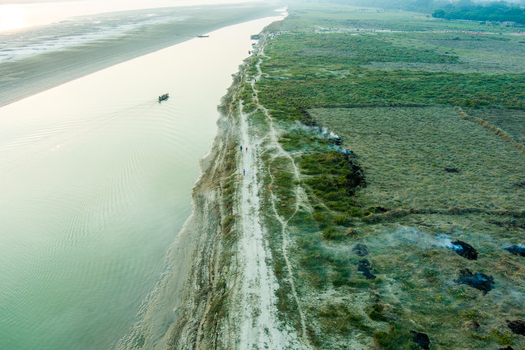 Aerial view of the riverbank with a small boat on the water a dirt path with people walking and green fields with patches of smoke in Rajshahi Rajshahi Division Bangladesh.