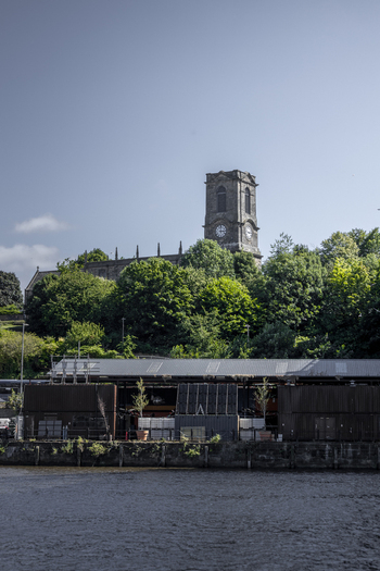 View of the historic stone church tower with a clock face rising above lush green trees and industrial riverside structures in Newcastle upon Tyne England United Kingdom. View of the historic stone church tower with a clock face rising above lush green trees and industrial riverside structures in Newcastle upon Tyne England United Kingdom.